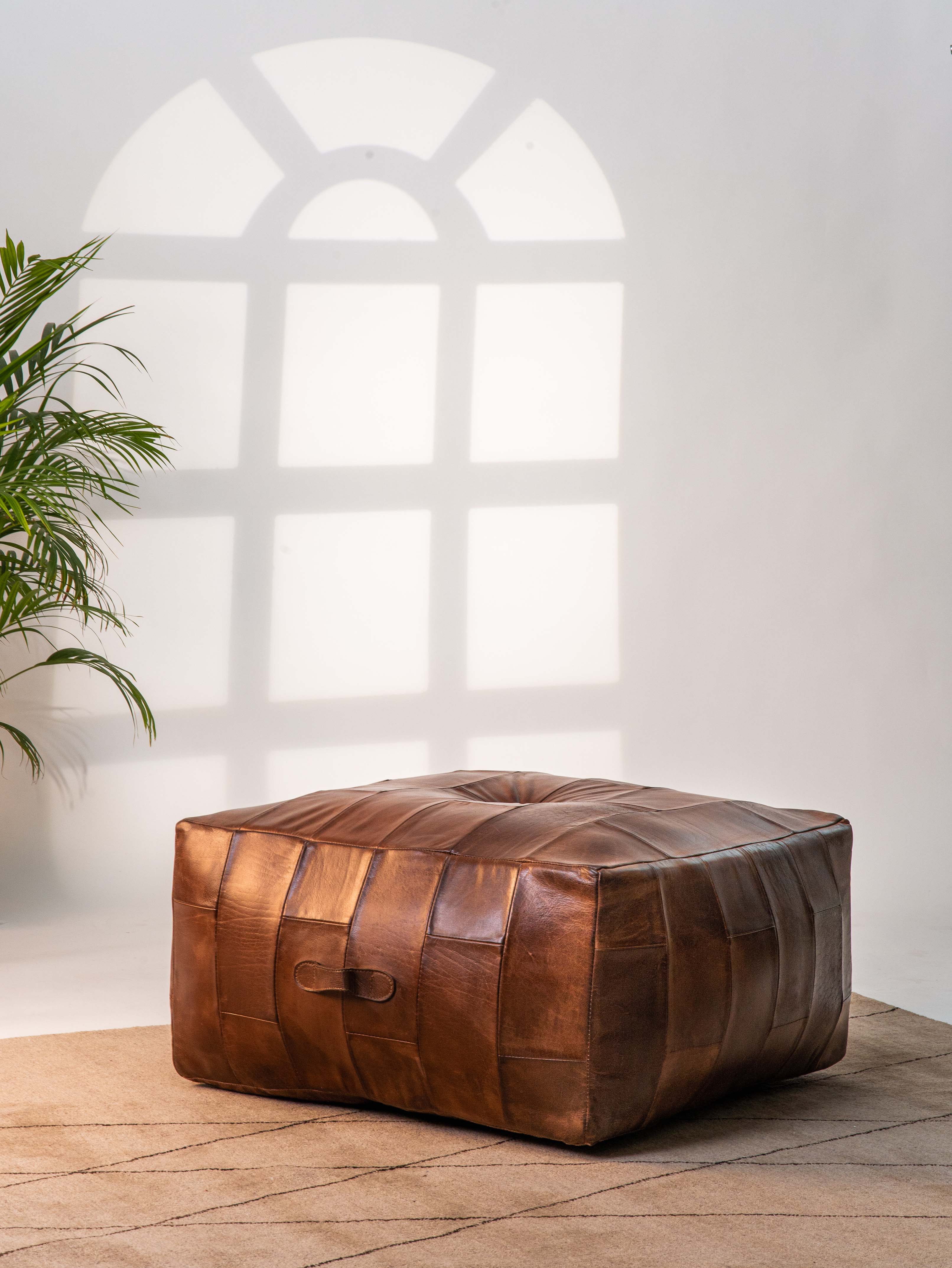 Square buffalo leather pouf in cognac color beside beige couch and indoor plant