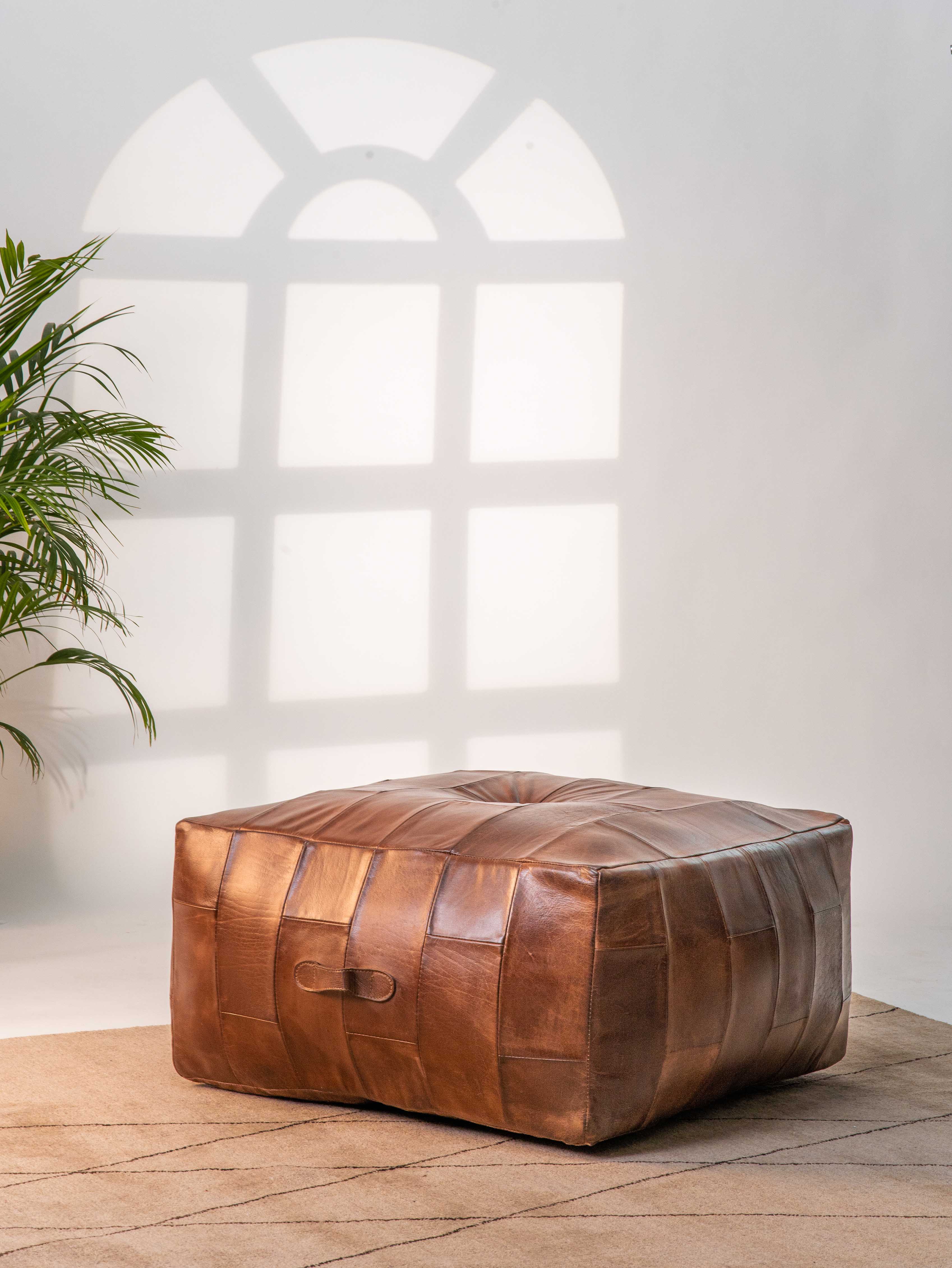 Square buffalo leather pouf in cognac color beside beige couch and indoor plant